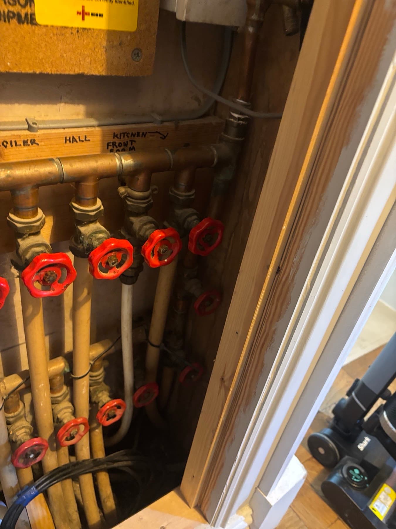Old copper underfloor heating manifold with red-wheel gate valves on every circuit, each labelled in pen on the wall