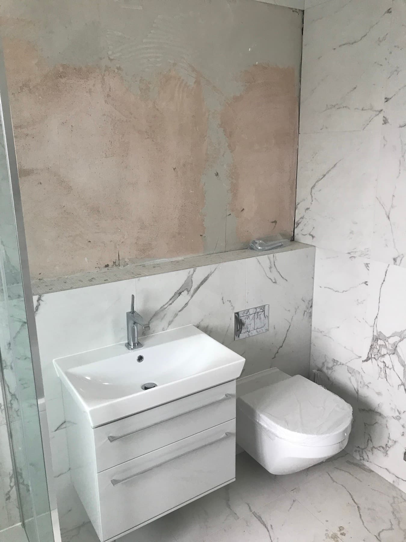 Wall-hung toilet and vanity basin on a marble-effect tiled wall, mid-job