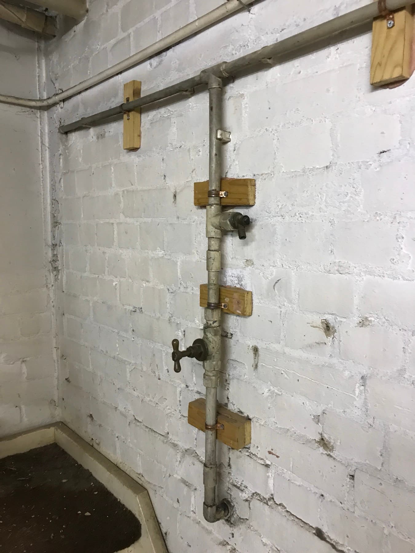 Old galvanised rising main with a stopcock, clipped to wooden battens on a painted brick wall